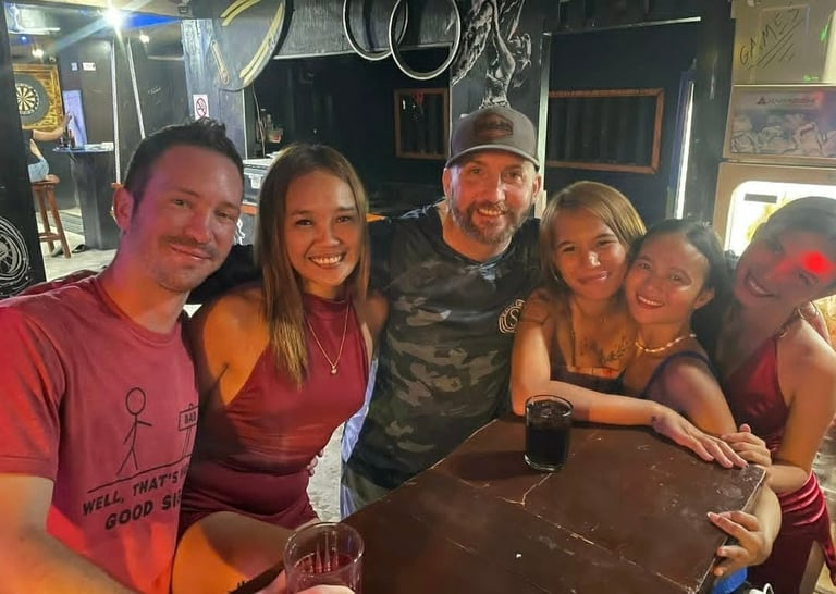 Six people posing together at a bar, smiling at the camera while standing around a dark counter with drinks