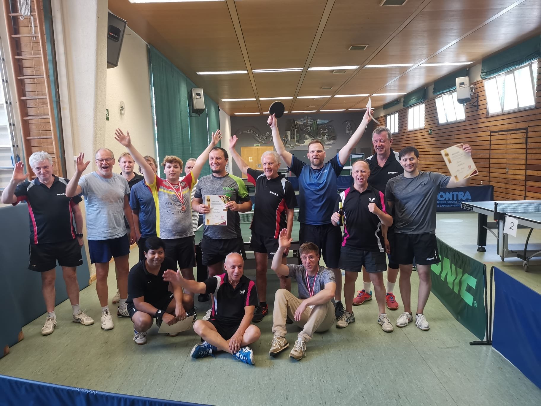 Group of sports team members posing with arms raised in indoor gymnasium, celebrating together with enthusiasm.