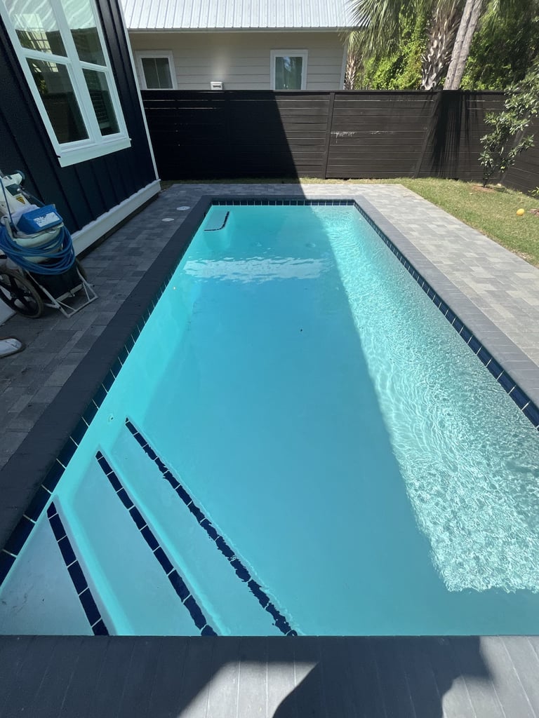 A bright turquoise rectangular swimming pool in a modern backyard with black tiles, concrete decking, and a dark-fenced perimeter next to a contemporary house
