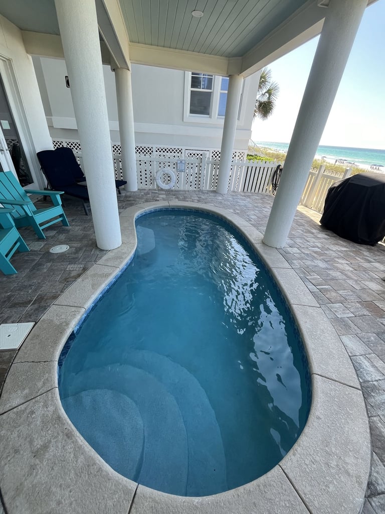Oval backyard pool with blue water on a covered patio overlooking the ocean and beach