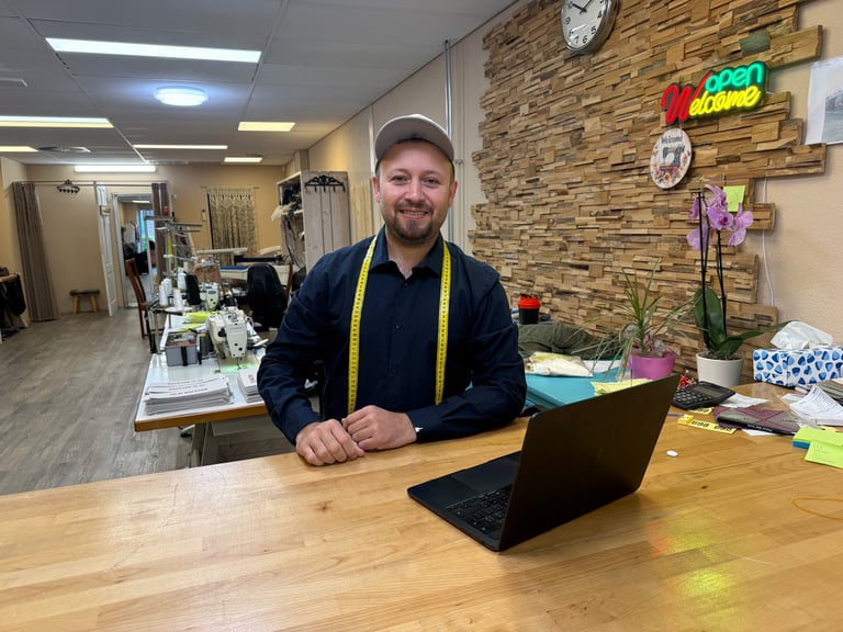 Man with beard wearing navy jacket and yellow lanyard sits at wooden desk with laptop in casual office space with brick wall and neon signs