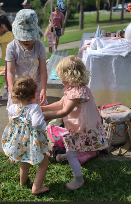 Young girl in floral dress examining items at an outdoor community yard sale or flea market on grass