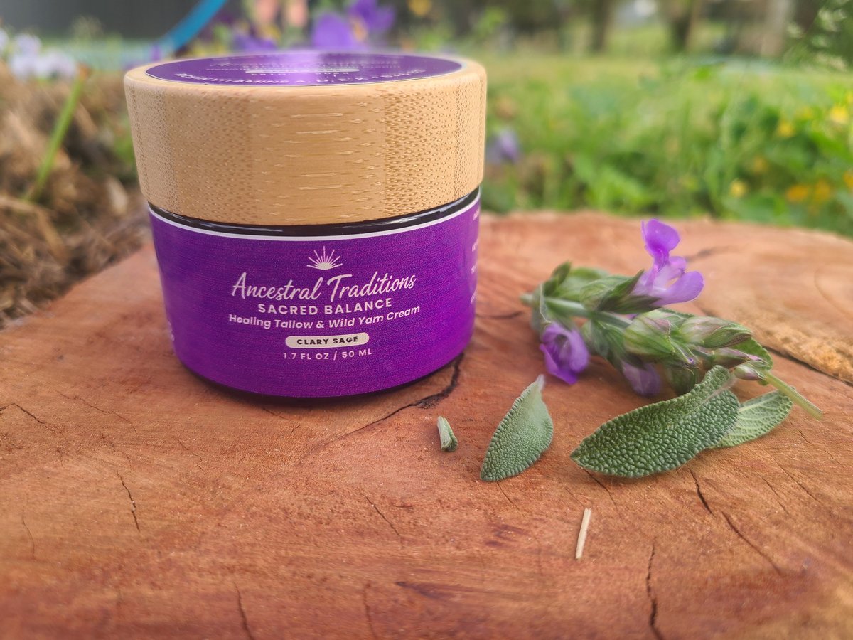 Purple jar of Ancestral Traditions Sacred Balance skincare product with wooden lid on wooden surface, surrounded by fresh herbs and purple flowers