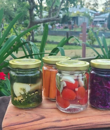 Four glass jars of preserved vegetables on a wooden surface in a garden setting, containing pickled onions, carrots, radishes, and beets