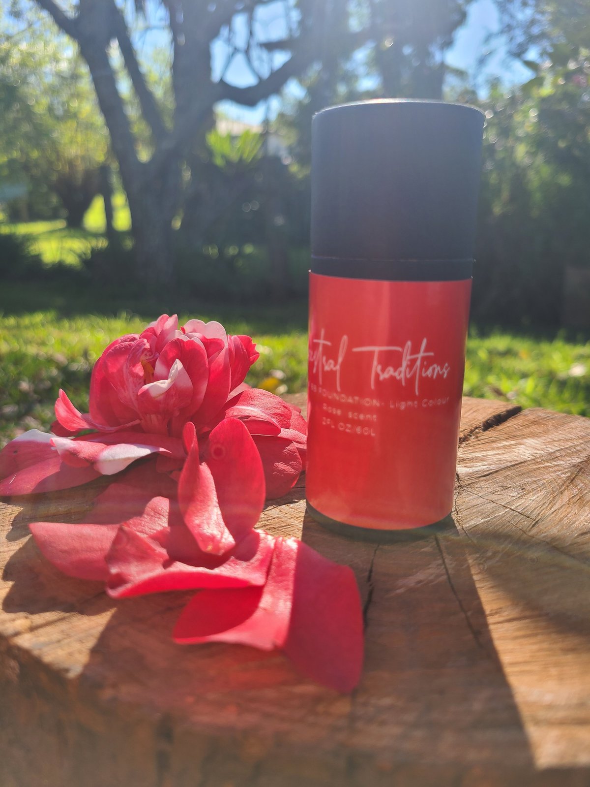 Red Ancestral Traditions BB Foundation Dark Colour cardboard tube bottle next to a pink rose on a wooden surface in a sunny garden
