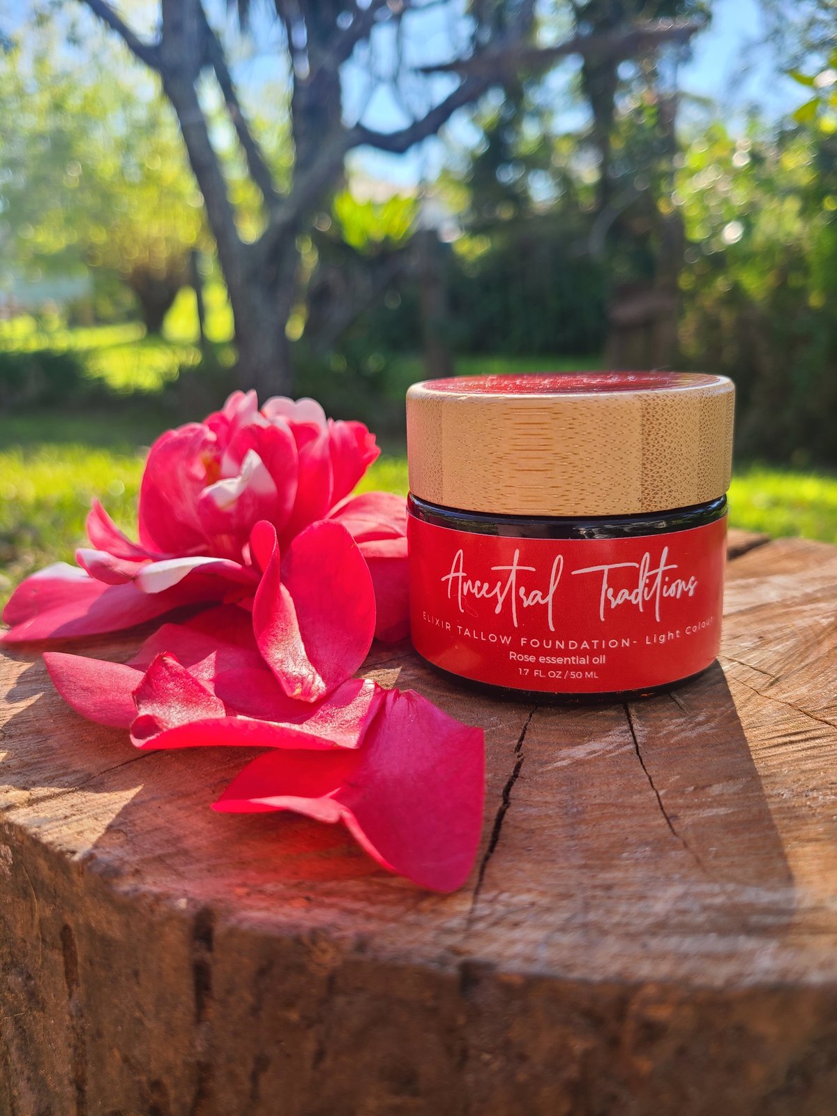 Red cosmetic jar with wooden lid labeled Ancestral Traditions Elixir Tallow Foundation Light Colour Rose essential oil, beside a pink peony flower on a wooden surface outdoors