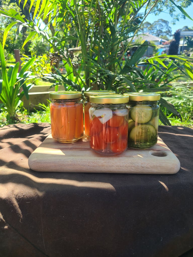 Four glass jars of preserved vegetables and pickled foods on a wooden board in a sunny garden setting