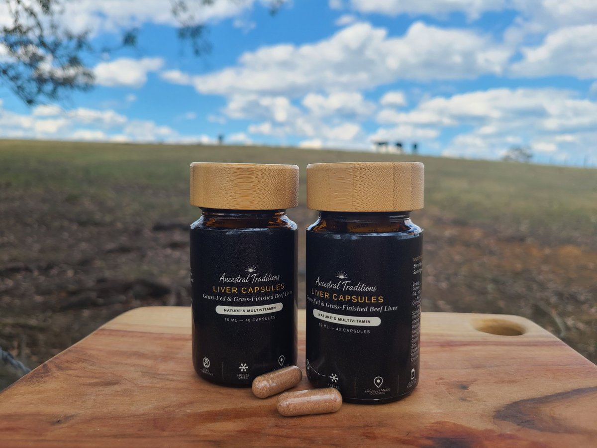 Two dark glass supplement bottles with wooden caps on a wooden surface outdoors, with pills nearby and open farmland in background