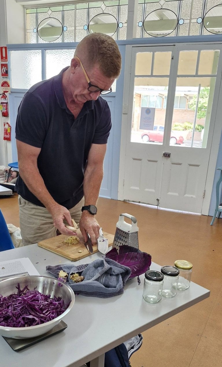 Man in dark polo shirt chopping red cabbage on a cutting board in a kitchen, with prepared vegetables and cooking utensils on the table