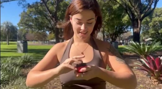 Woman in park holding red fruit, smiling while looking down at her hands with trees and green lawn in background
