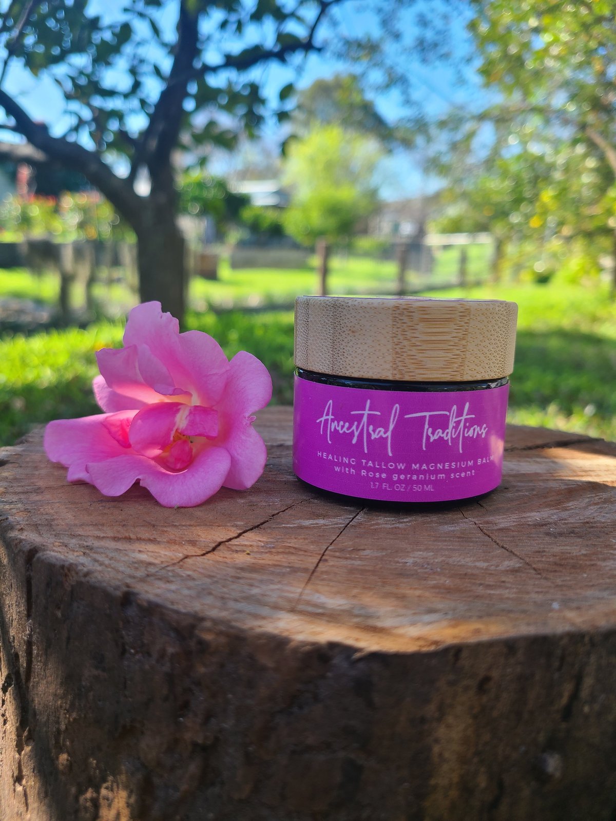 Pink Ancestral Traditions Healing Tallow Magnesium Balm with rose geranium scent beside a pink flower on a wooden tree stump in a sunny garden