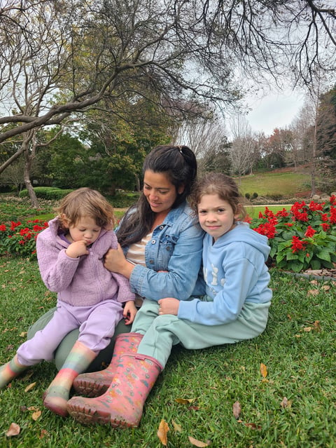 Woman and two girls sitting together on grass in a park with red flowers and trees in the background
