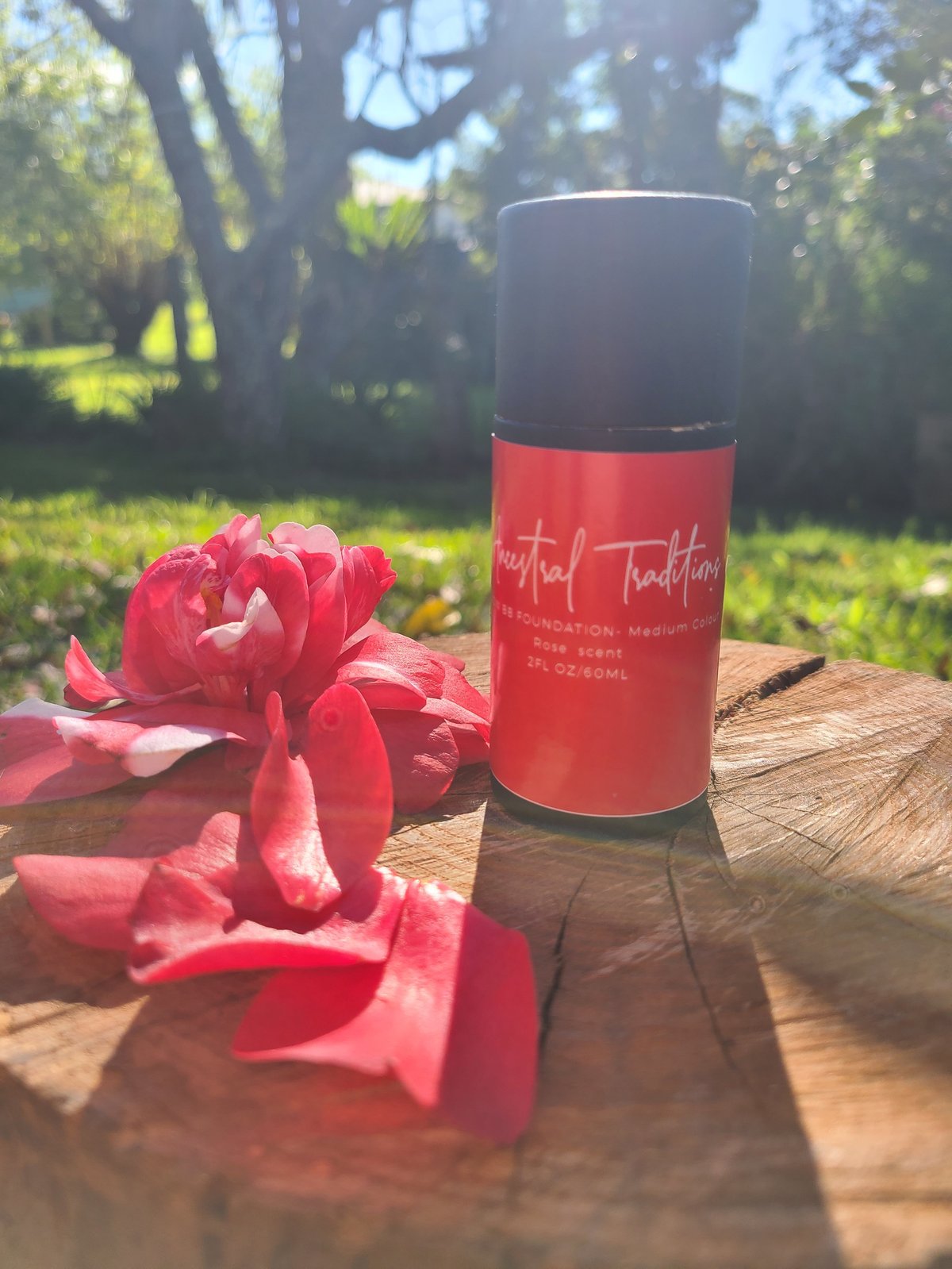 Red Ancestral Traditions BB Foundation Medium Colour cardboard tube bottle next to fresh red flowers on a wooden surface in a sunlit garden