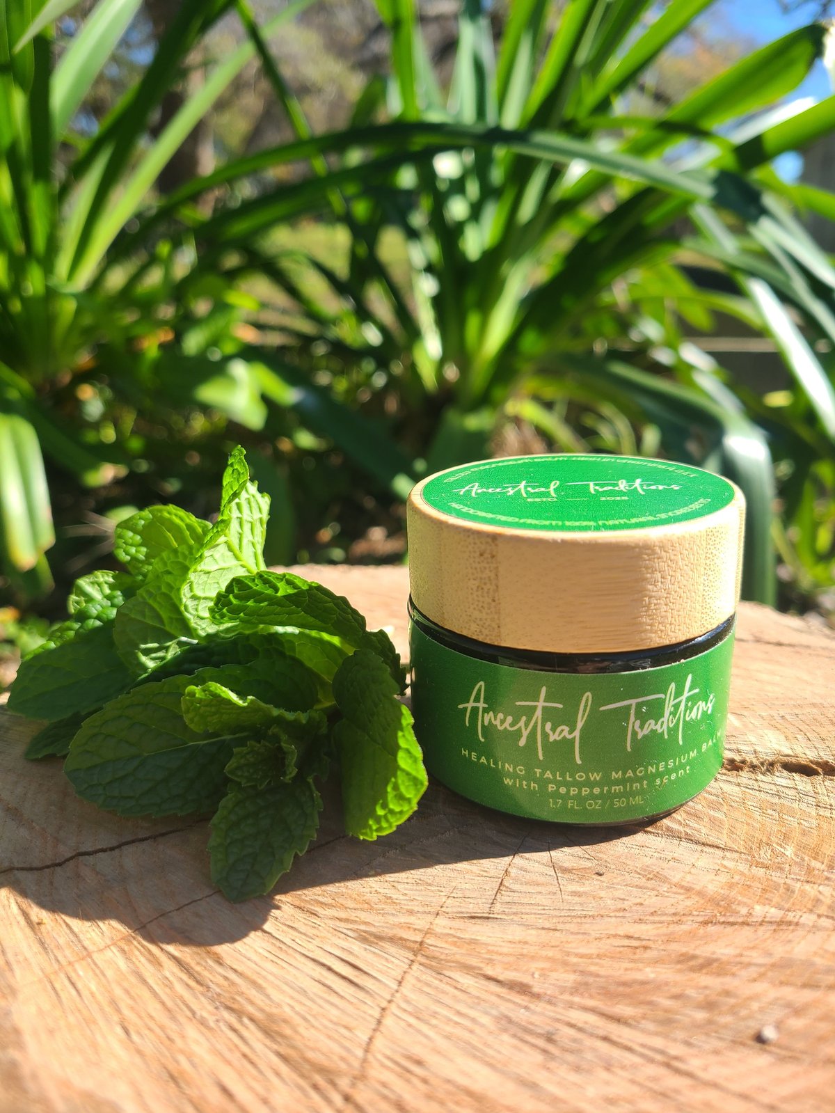 Green Ancestral Traditions Healing Tallow Magnesium Balm with peppermint scent next to fresh mint leaves on wooden surface with tropical plants in background