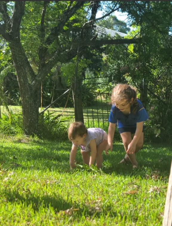 Adult and child playing outdoors on grass beneath large trees with fence and house visible in background