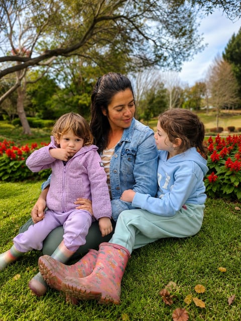 Woman with two young children sitting on grass in a park surrounded by red flowers and green trees