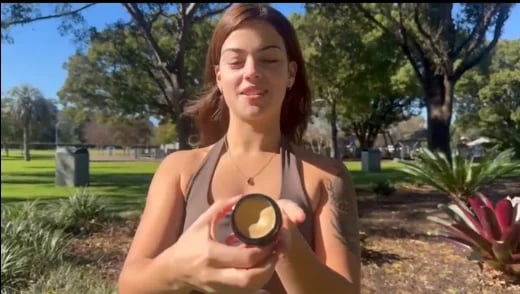Woman in halter top holding a glass jar with amber liquid outdoors in a park with grass and trees