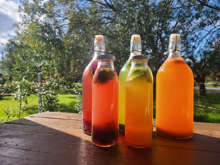 Three glass bottles filled with colorful beverages - red, yellow-green, and orange - on a wooden table with green garden and trees in background