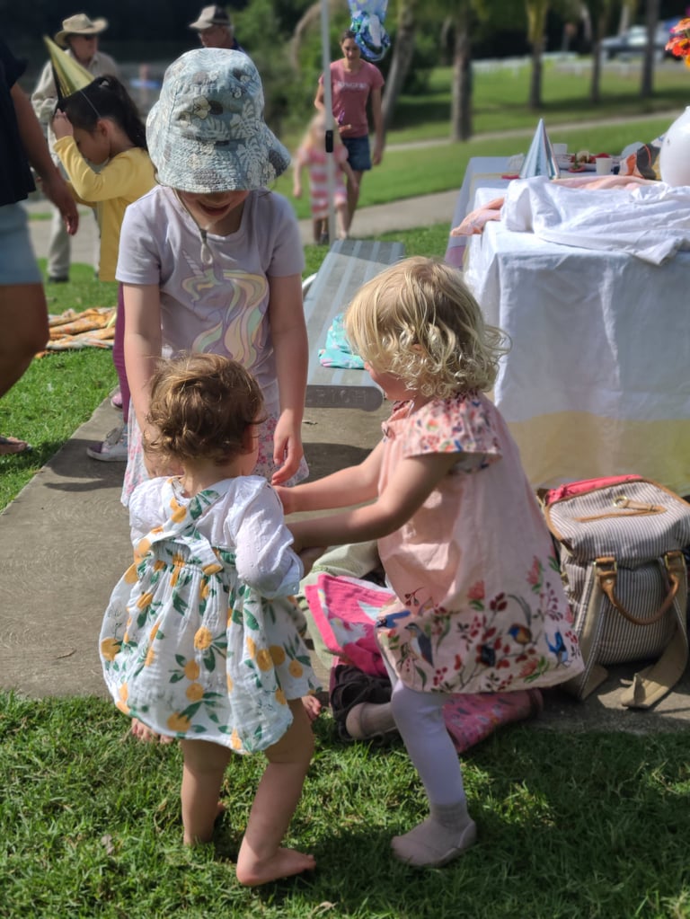 Young woman in floral dress and sun hat with two toddlers at an outdoor community event in a park