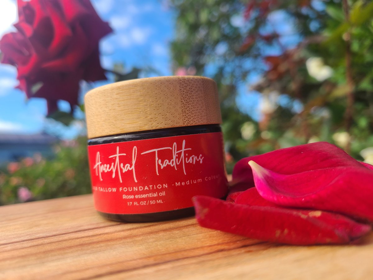 Red Ancestral Traditions tallow BB foundation jar in Medium Colour with rose essential oil, surrounded by red rose petals on a wooden surface in a garden setting