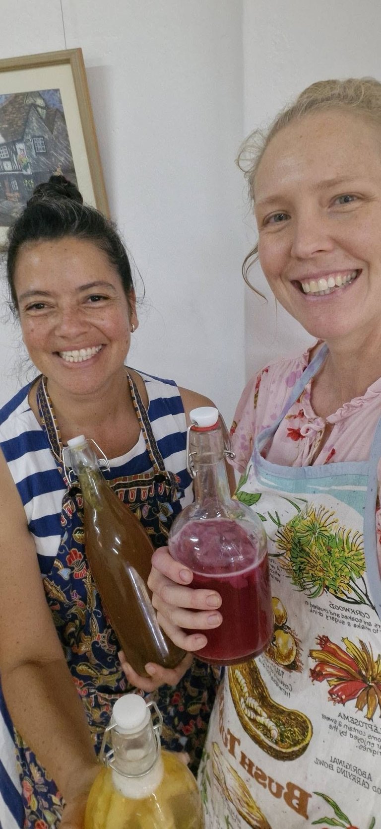 Two women smiling at camera, holding homemade fermented beverages in jars, wearing aprons in a kitchen setting