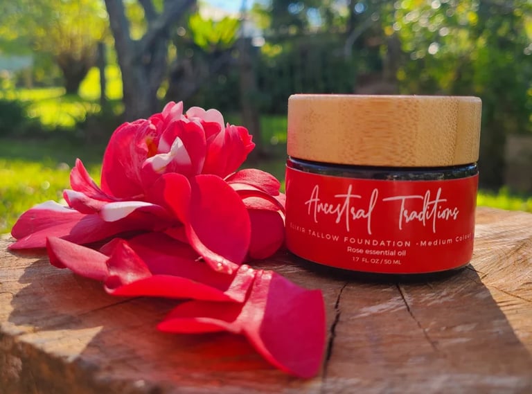 Red glass jar with wooden lid labeled Ancestral Traditions Elixir Tallow Foundation Medium Colour Rose essential oil, beside a red rose flower on a wooden surface outdoors