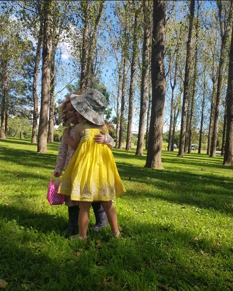 Woman in yellow dress and purple shoes wearing a hat, standing in a tree-lined park on green grass