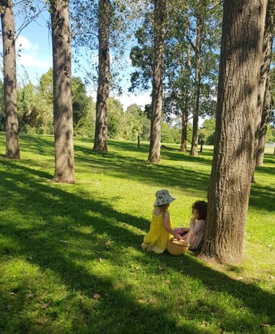 Two children sitting on grass between trees in a sunlit park with green lawn