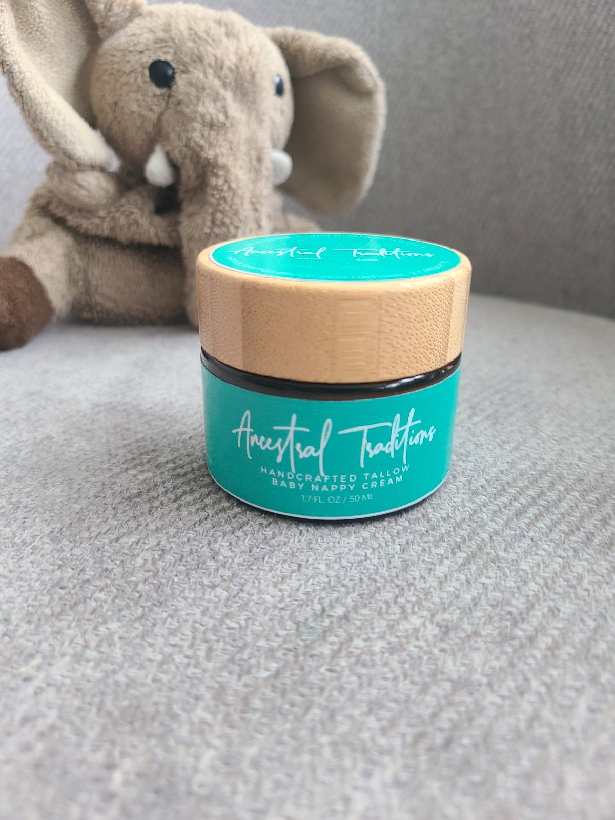 A turquoise jar of Ancestral Traditions handcrafted candle with wooden lid next to a plush elephant toy
