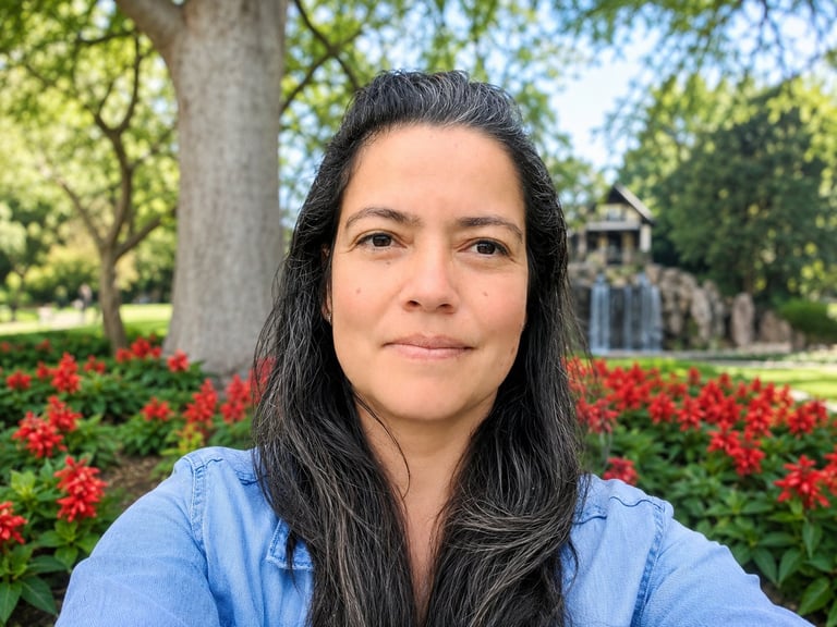 Woman with long dark hair wearing a light blue shirt, smiling at camera outdoors with red flowers and trees in background