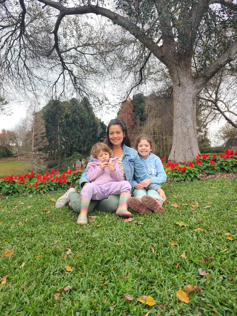 Woman with two young children sitting on grass under a large tree, with red flowers and autumn scenery in background