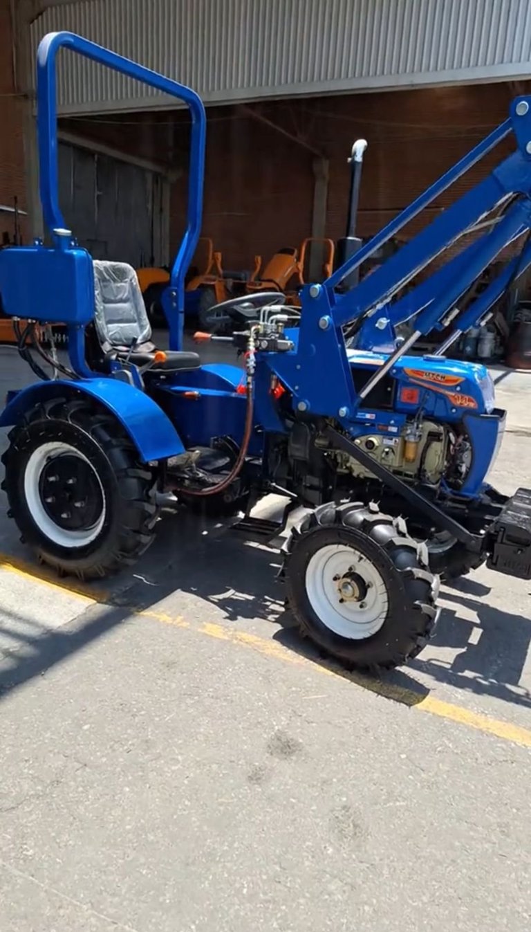 Bright blue compact tractor with attached farm equipment in front of a metal shed warehouse