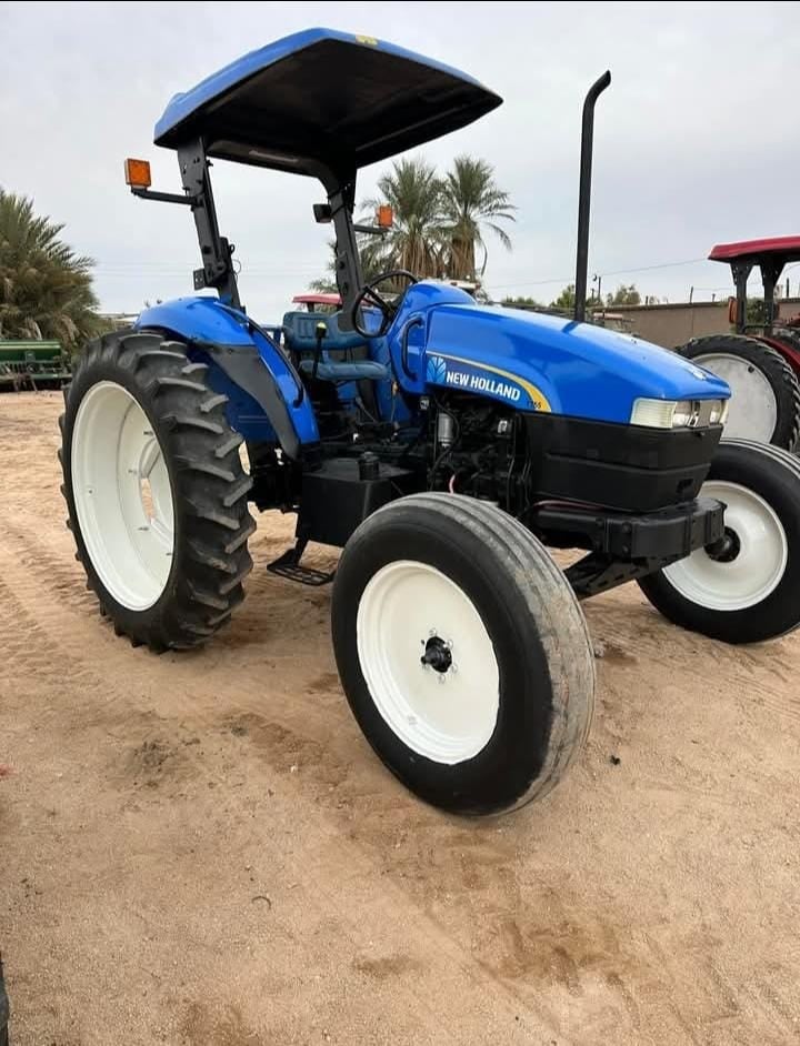 Blue New Holland utility tractor with canopy and white-wall tires parked in sandy lot with palm trees in background