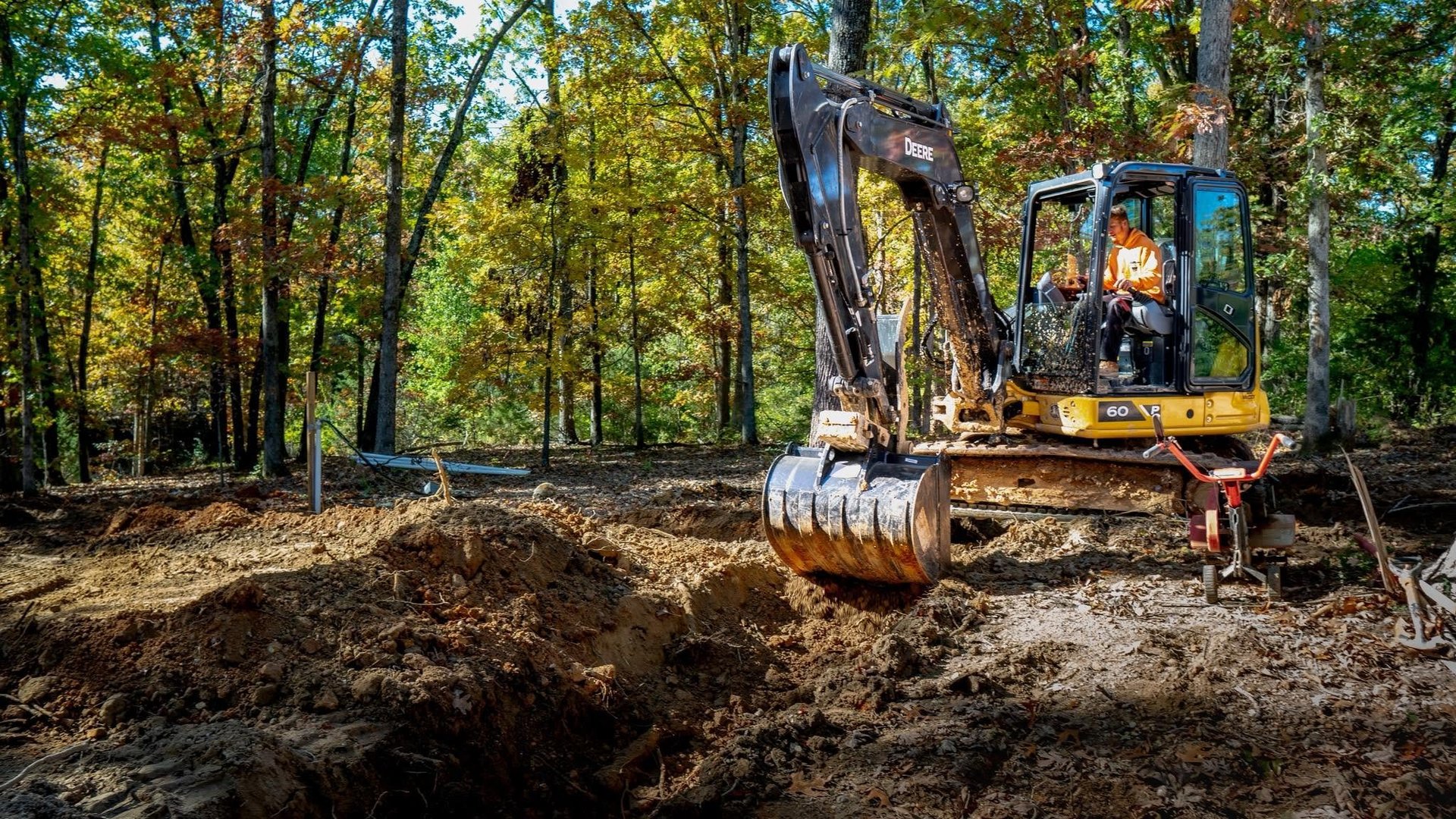 Yellow excavator with operator in cab digging in wooded area surrounded by trees on a sunny day