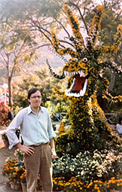Man standing next to large yellow dragon topiary with open mouth in garden setting surrounded by flowers and trees