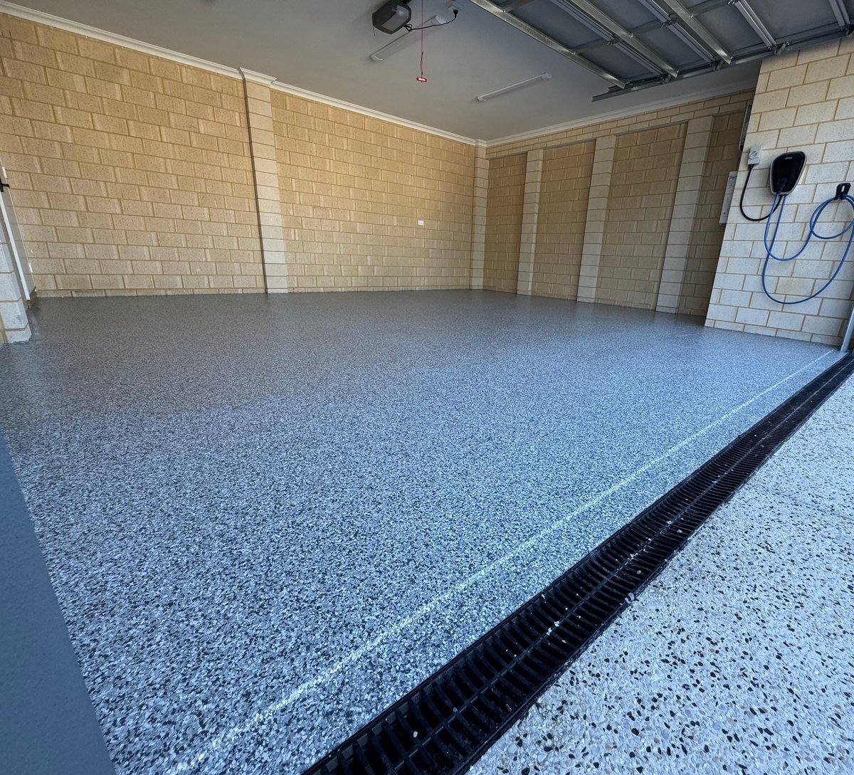 Empty garage with a fresh light grey and black speckled flake floor, brick walls and black drainage channel