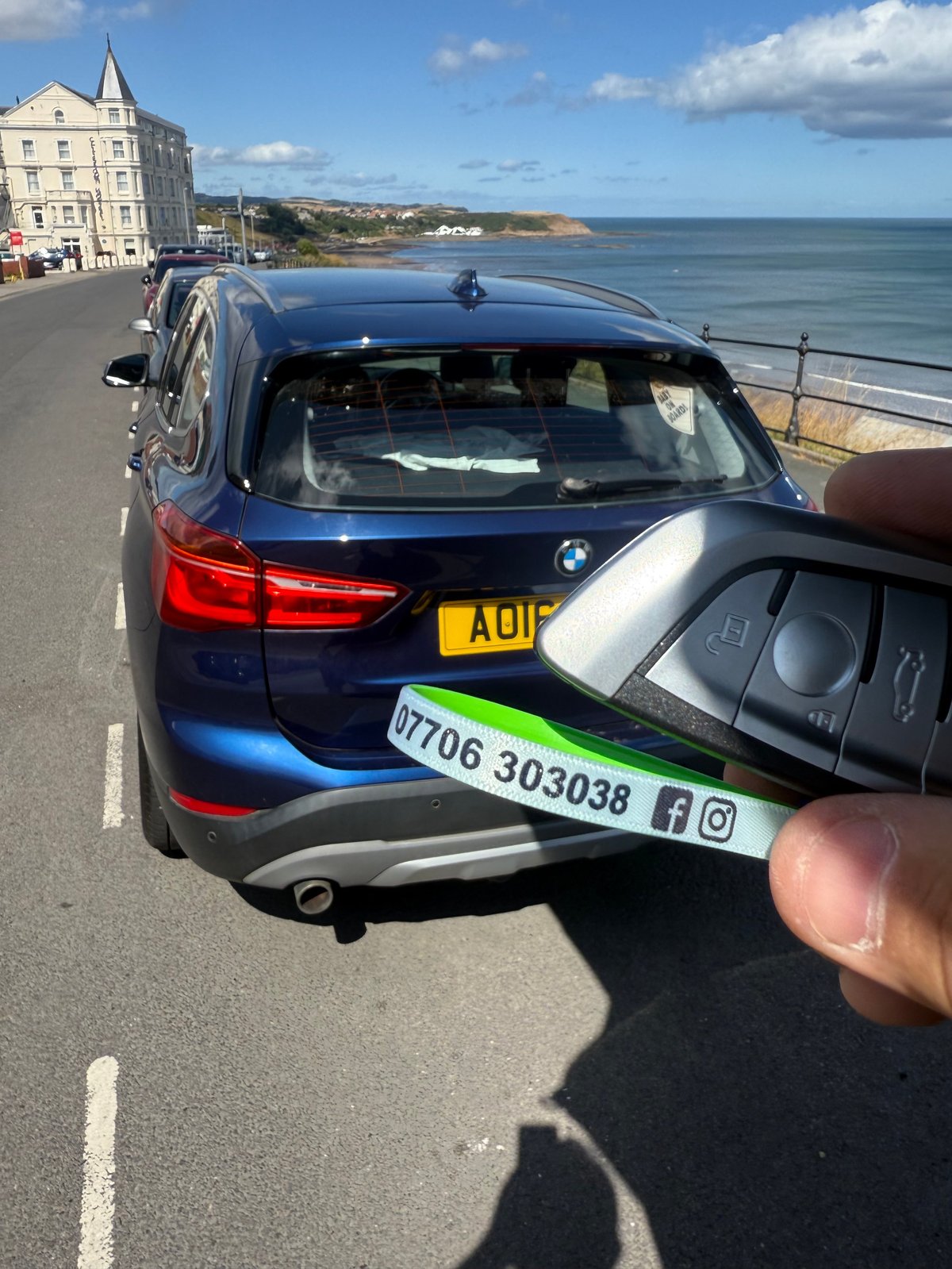 Blue BMW X1 parked on a seafront road on the South Cliff in Scarborough, with cliff-top hotels in the background and the North Bay behind. A fresh BMW smart key is held in the foreground with a green Scarborough Auto Locksmith lanyard showing 07706 303038 and Facebook/Instagram icons.