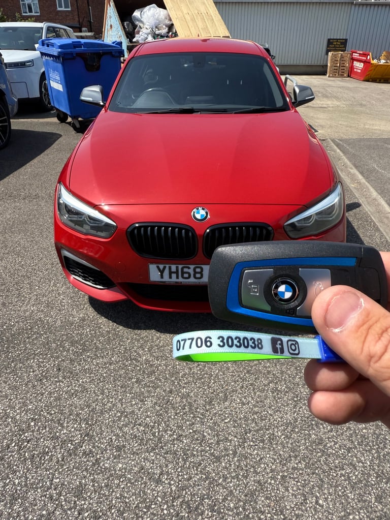 Hand holding a blue key fob with phone number in front of a red BMW car in a parking lot