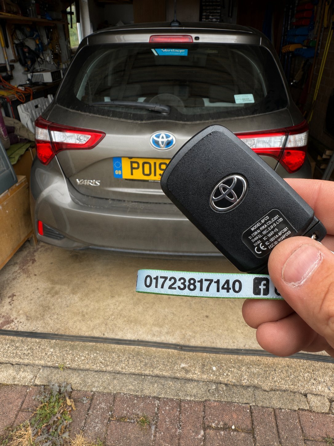 Hand holding a Toyota car key fob in front of a parked silver Toyota Yaris hybrid with UK license plate