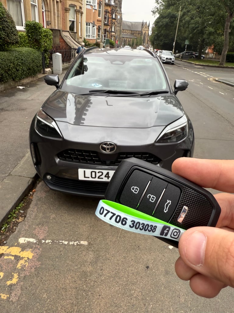 Hand holding black Toyota car key fob with contact details in front of parked silver Toyota Yaris on residential street