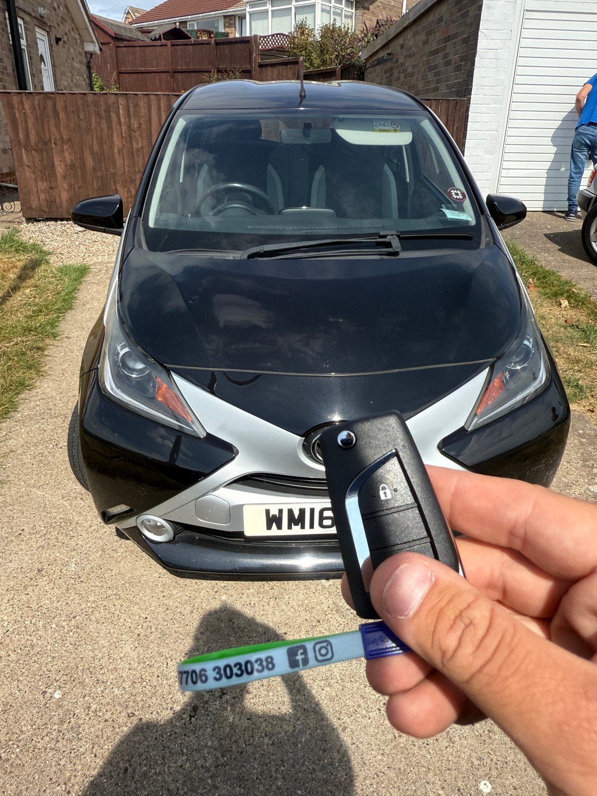Hand holding a Toyota key fob in front of a black Toyota Aygo city car parked in a residential driveway
