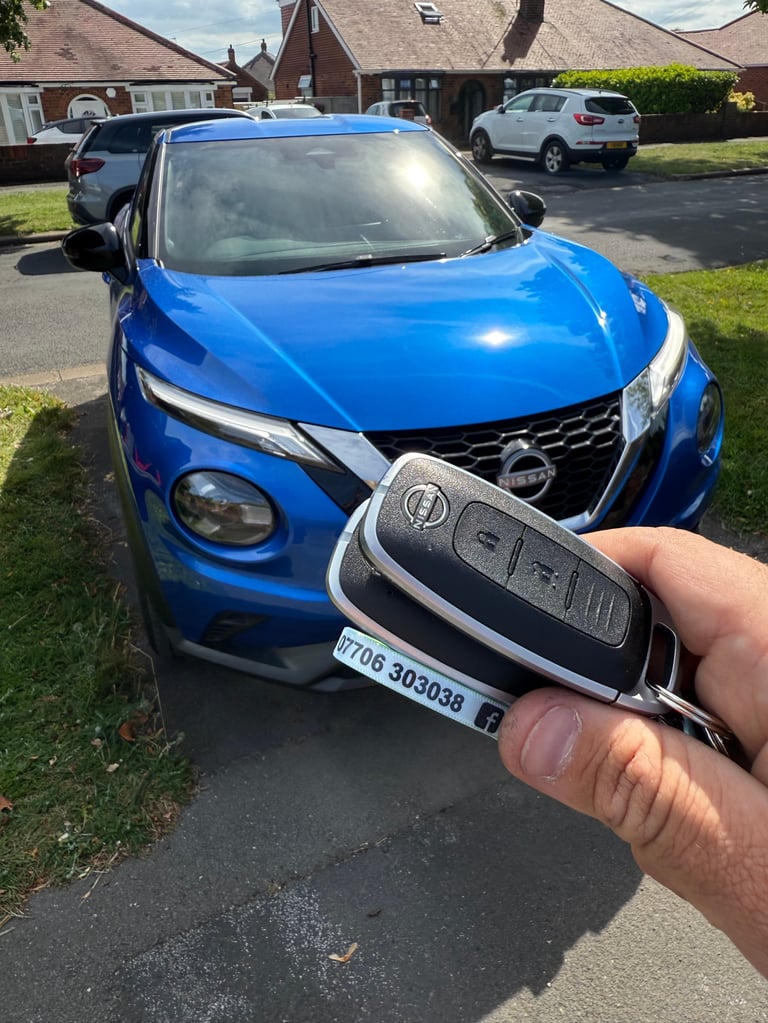 Hand holding a car key fob in front of a blue Nissan Juke parked in a residential driveway