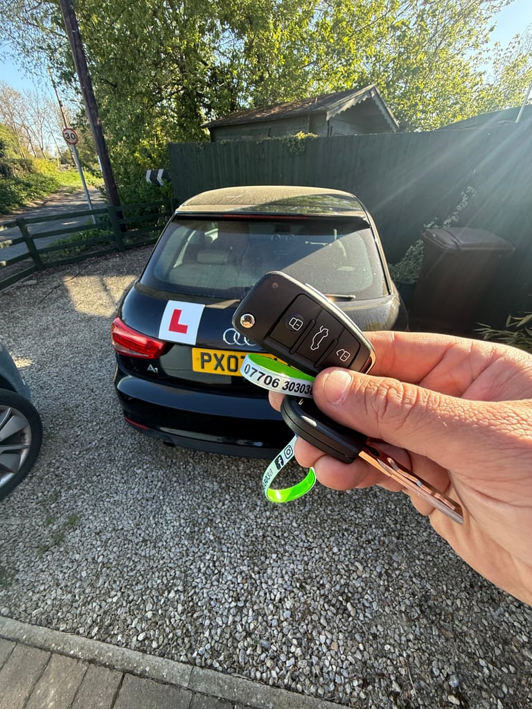 Hand holding a car key fob in front of a dark blue learner car with L-plates in a driveway