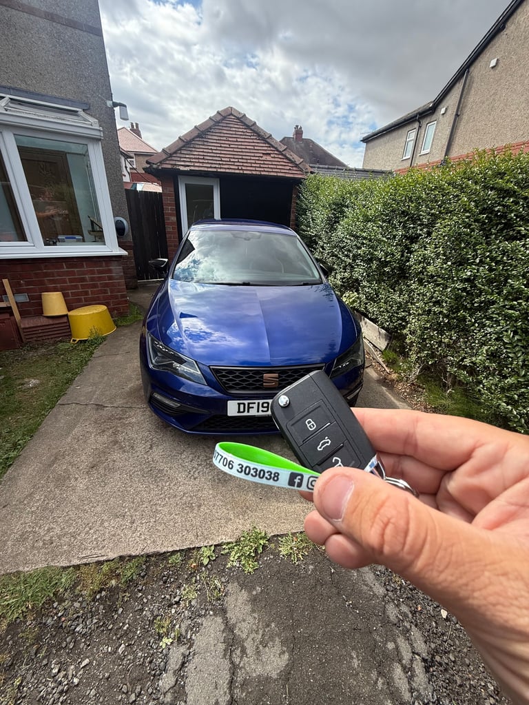 Hand holding car key fob in front of blue SEAT Leon parked in residential driveway with hedging behind