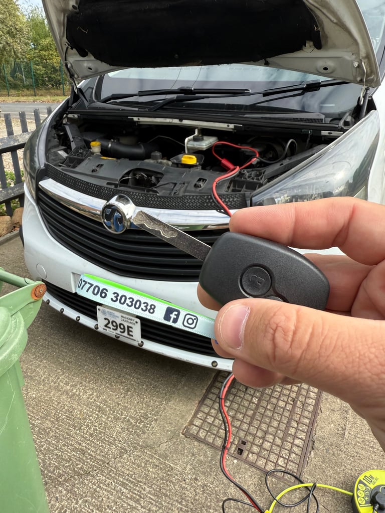 Hand holding a car key fob in front of an open white Vauxhall vehicle with engine bay visible