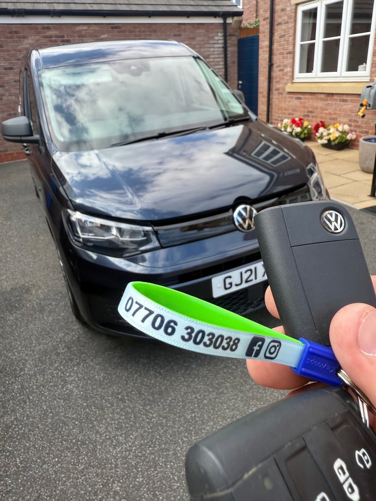 Hand holding Volkswagen car key fob with green wristband in front of black VW Caddy van parked outside brick house