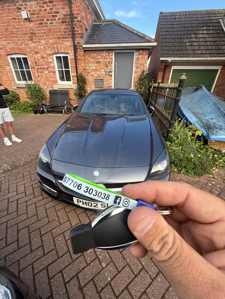 Hand holding a car key fob in a driveway with a gray BMW sedan parked behind, red brick houses visible