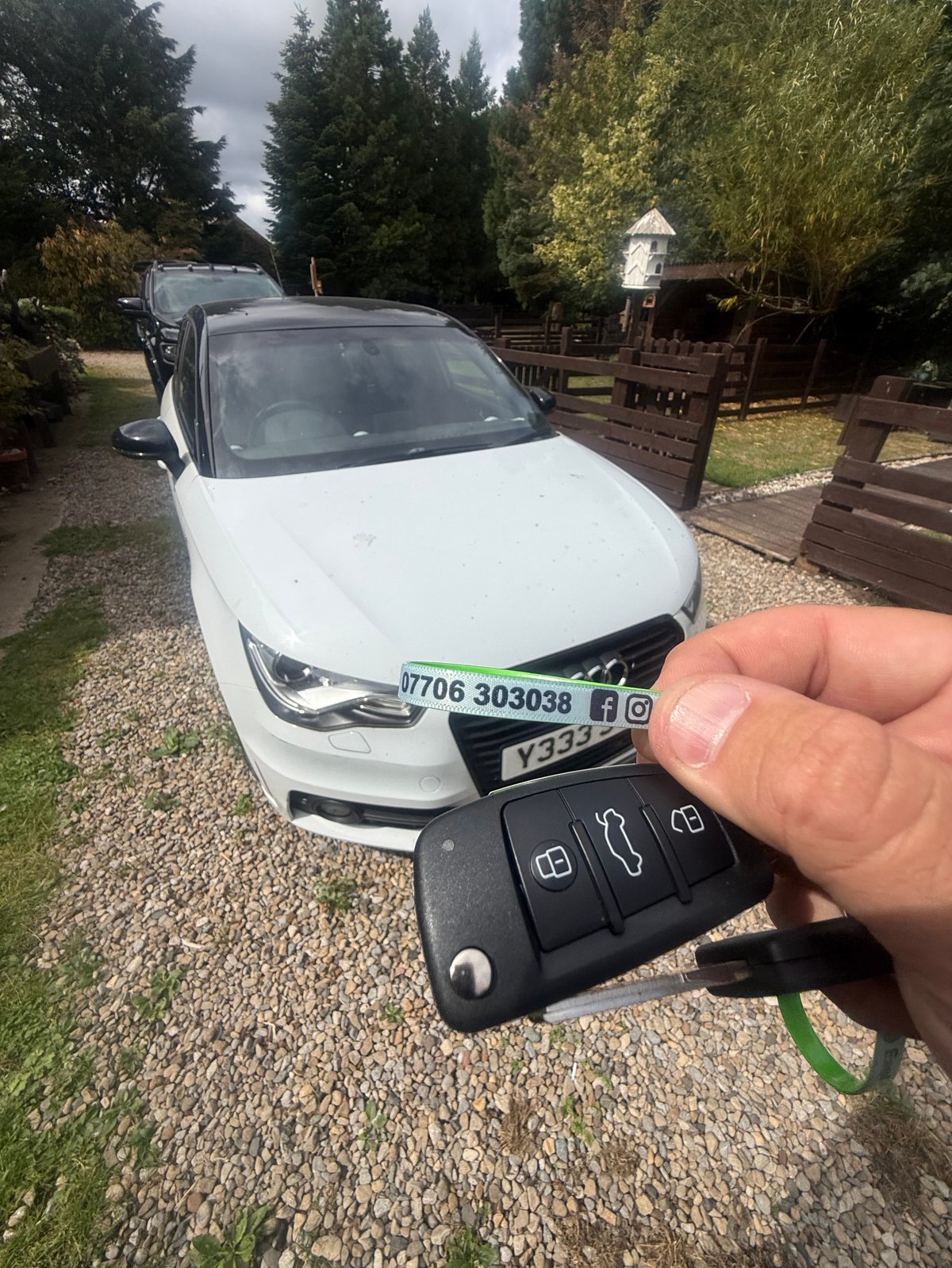 Hand holding car key fob in front of white Audi A1 sedan parked in gravel driveway with trees and birdhouse in background