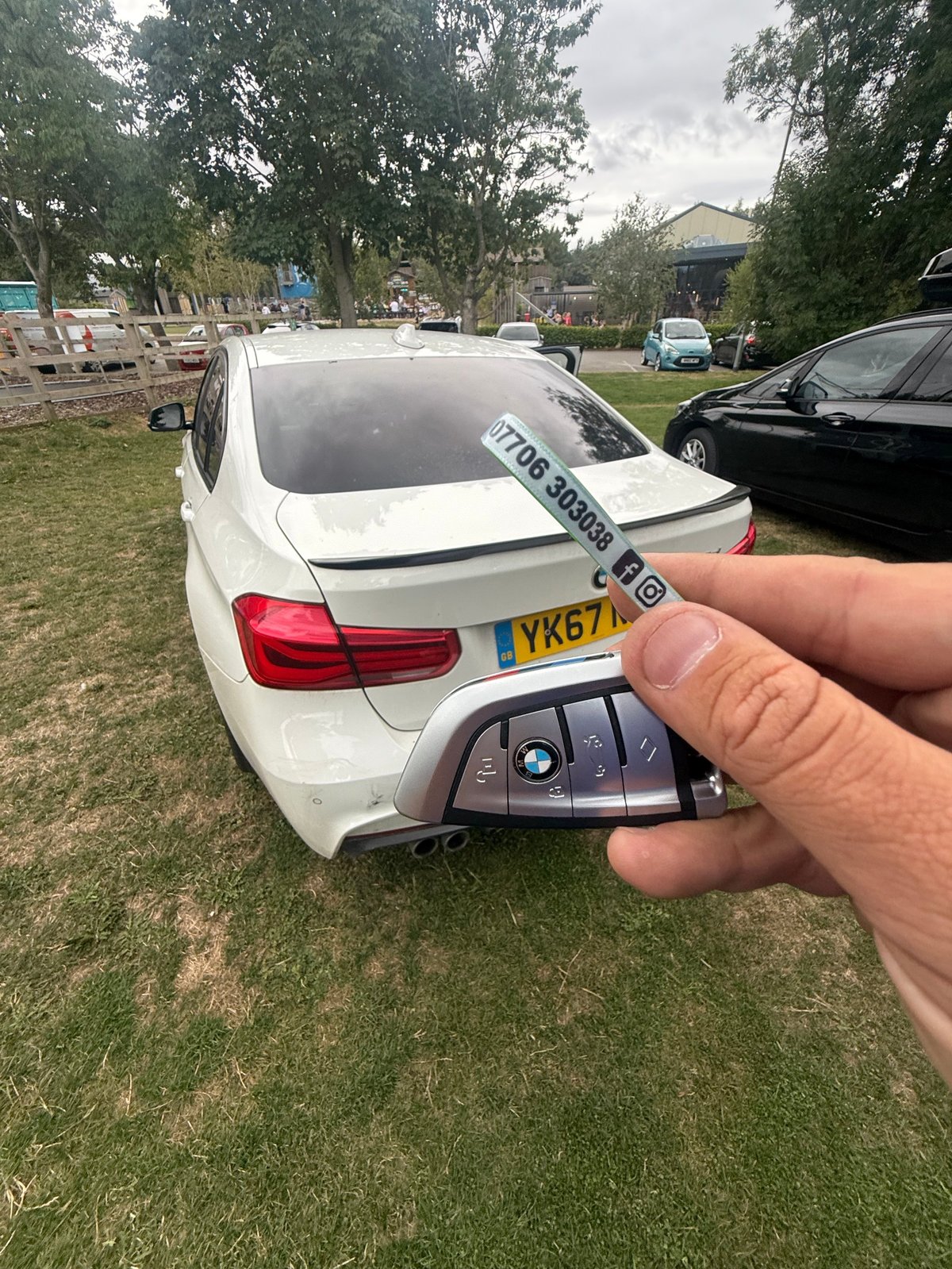 Person holding car key fob in front of white BMW sedan parked on grass with trees in background at a Scarborough event