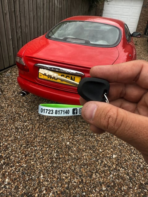 Red 1999 Jaguar XKR coupe parked on a gravel driveway in Brandesburton, new key fob held in the foreground with Scarborough Auto Locksmith lanyard showing 01723 817140